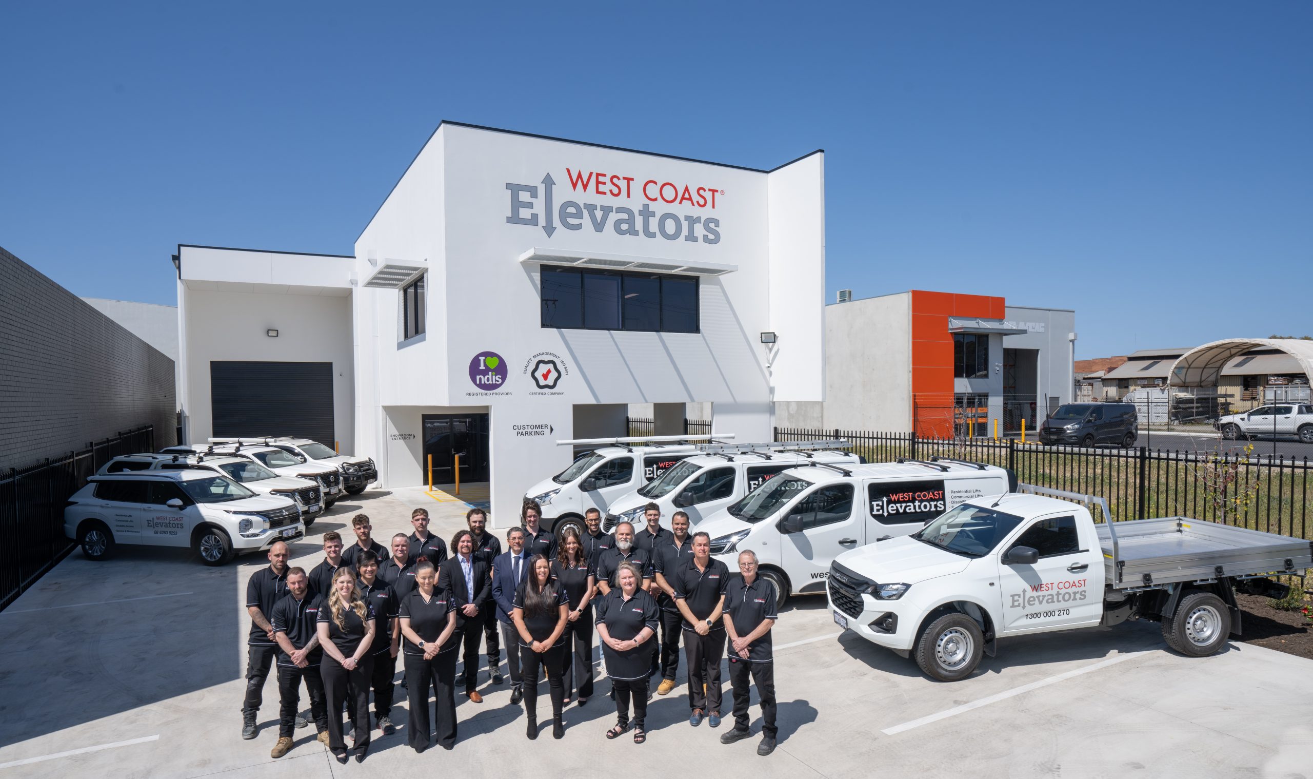 A group of people wearing matching black polo shirts stands in front of a building with the logo "West Coast Elevators" prominently displayed. Several white company vehicles are parked nearby, all branded with the same logo. The building features additional logos, including one that says "I love NDIS." The sky is clear and blue, suggesting a bright, sunny day.