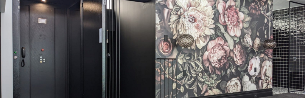 A modern, dark-colored elevator with an open door on the left. On the right, a wall is decorated with large, detailed floral wallpaper featuring roses and peonies in muted tones. There is a spherical, woven light fixture on the wall. The background features a tiled area with reflective, black tiles.