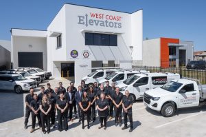 A group of people wearing black uniforms is standing together in front of a building with the sign "West Coast Elevators" on it. Several white company vehicles with the same branding are parked to the side. The building has logos indicating it is an NDIS registered provider and a certified company for quality management systems. The scene is set under a clear blue sky.