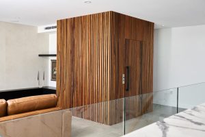 A modern interior features a stylish wooden panelled lift with vertical strips in a corner. Adjacent to the lift is a brown leather sofa. A partially visible wall-mounted piece of art is near the lift. Two tall black and white candles are displayed on a nearby ledge. The flooring is a mix of light beige tiles and marble. Glass panels can be seen on the right, suggesting a staircase or landing area, and the ceiling is kept minimal with a white, smooth finish and recessed lighting fixtures.