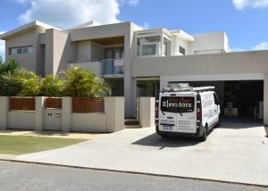 Modern two-storey house with a white and light grey facade, surrounded by a white and wooden fence. A van, labelled "West Coast Elevators," is parked in the driveway. There are palm trees in front of the house, and the garage door is open.